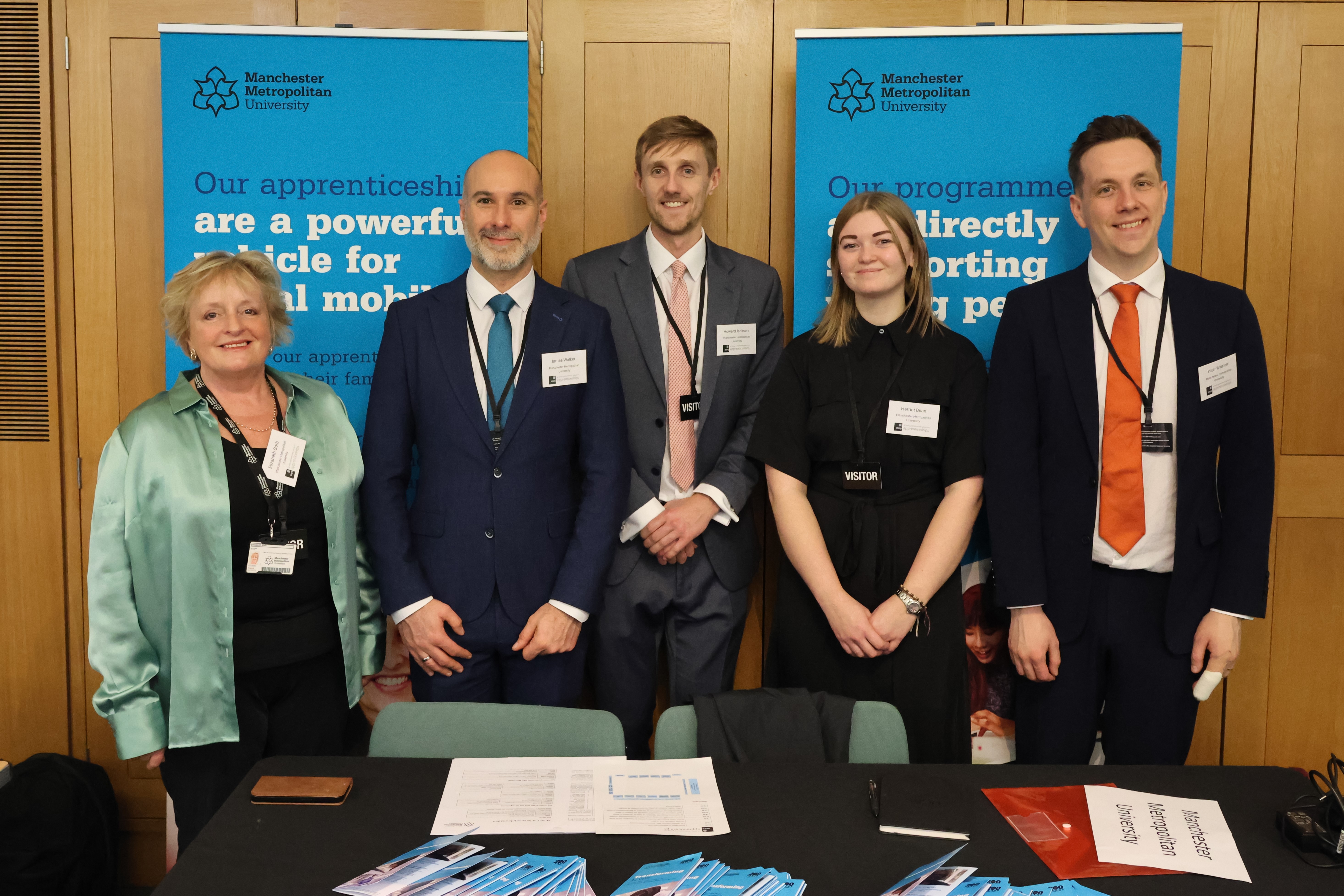 A group of Manchester Met Staff and Apprentices at the All-Party Parliamentary Group Apprenticeships Fair held at the House of Commons. Stood around a table with Manchester Met banners and marketing collateral